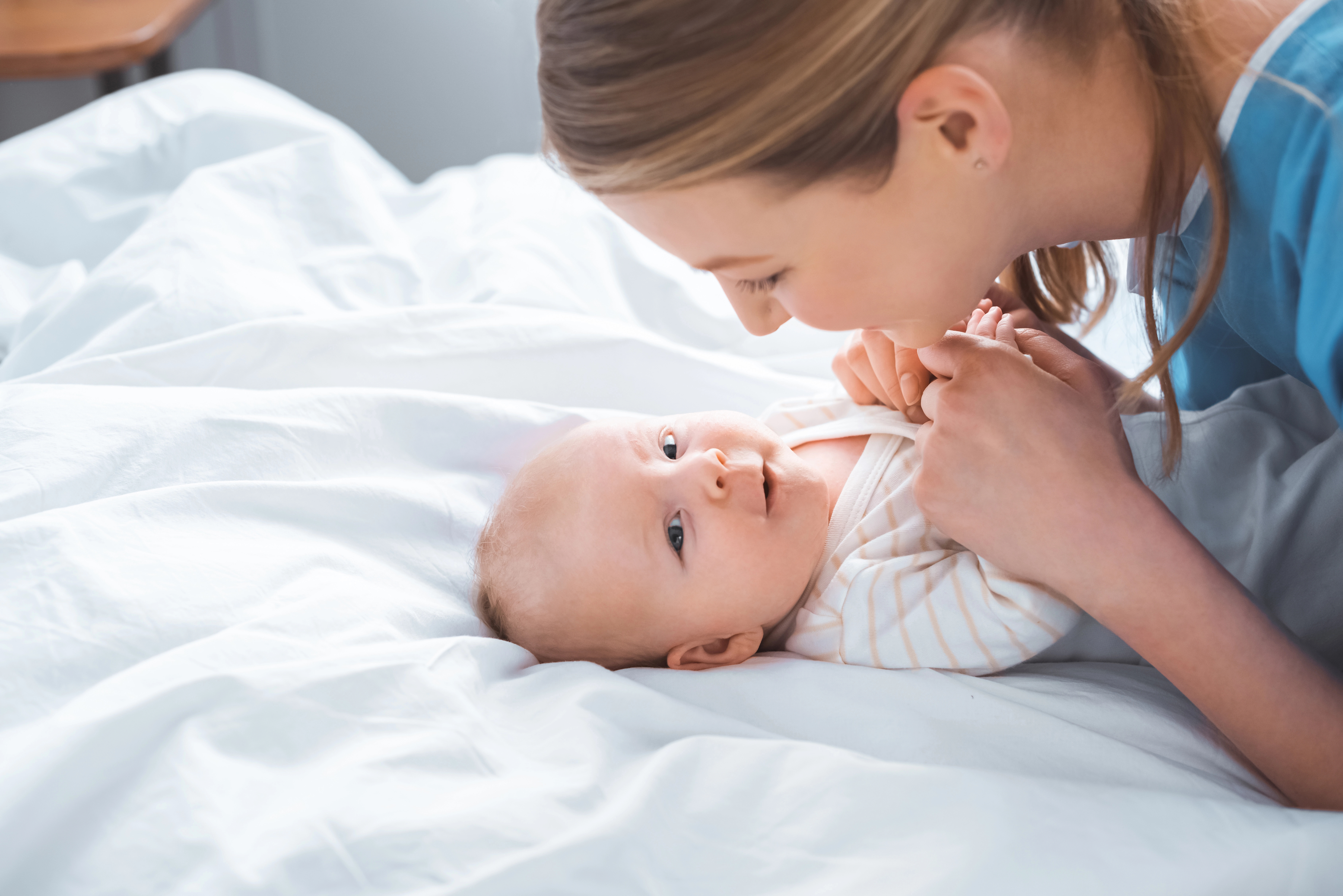 A woman smiles at a baby lying on a bed, holding its hands. The baby looks up, dressed in a striped outfit, surrounded by white bedding.