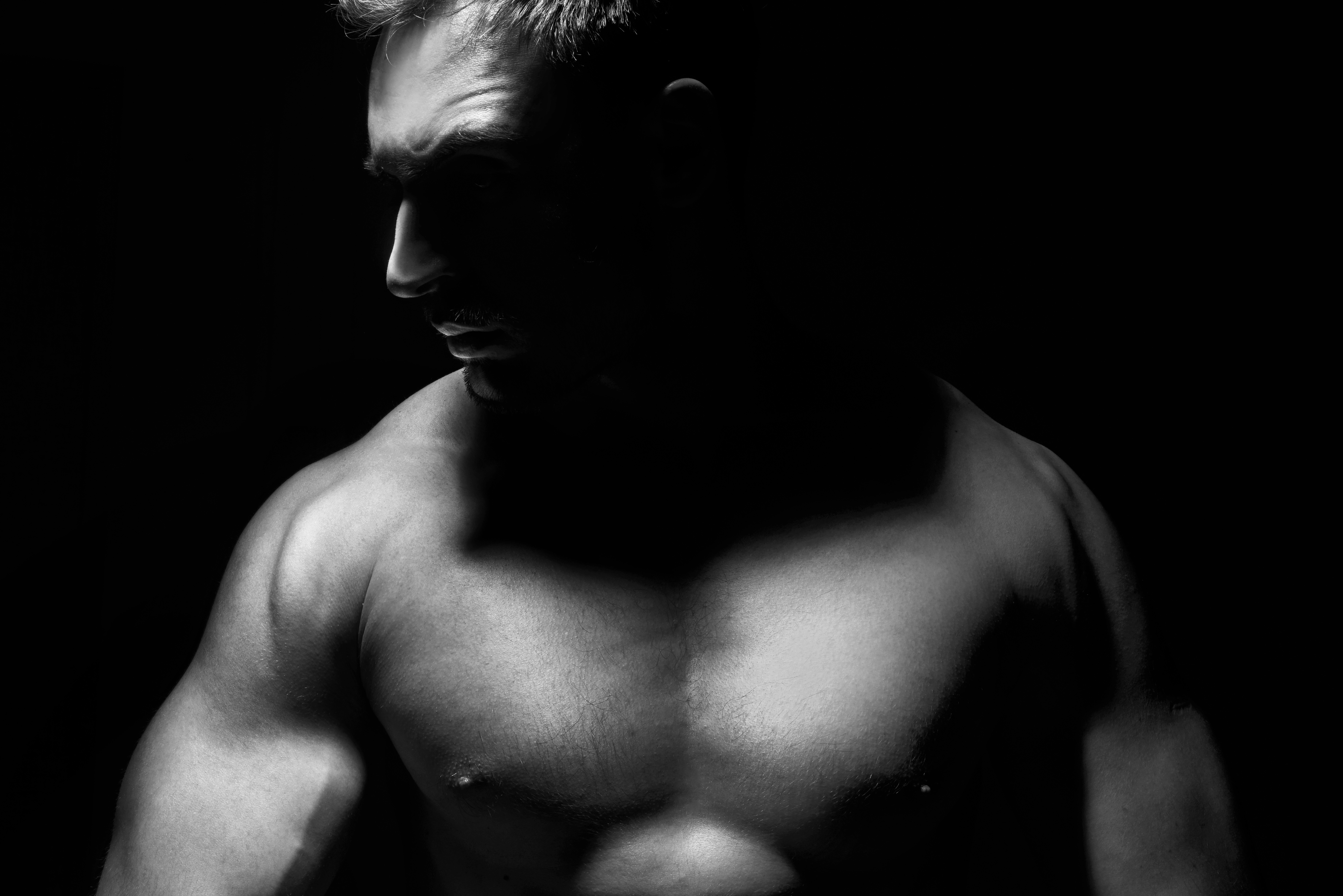 A muscular man is shown in dramatic lighting, highlighting his toned upper body and casting shadows on his face against a dark background.