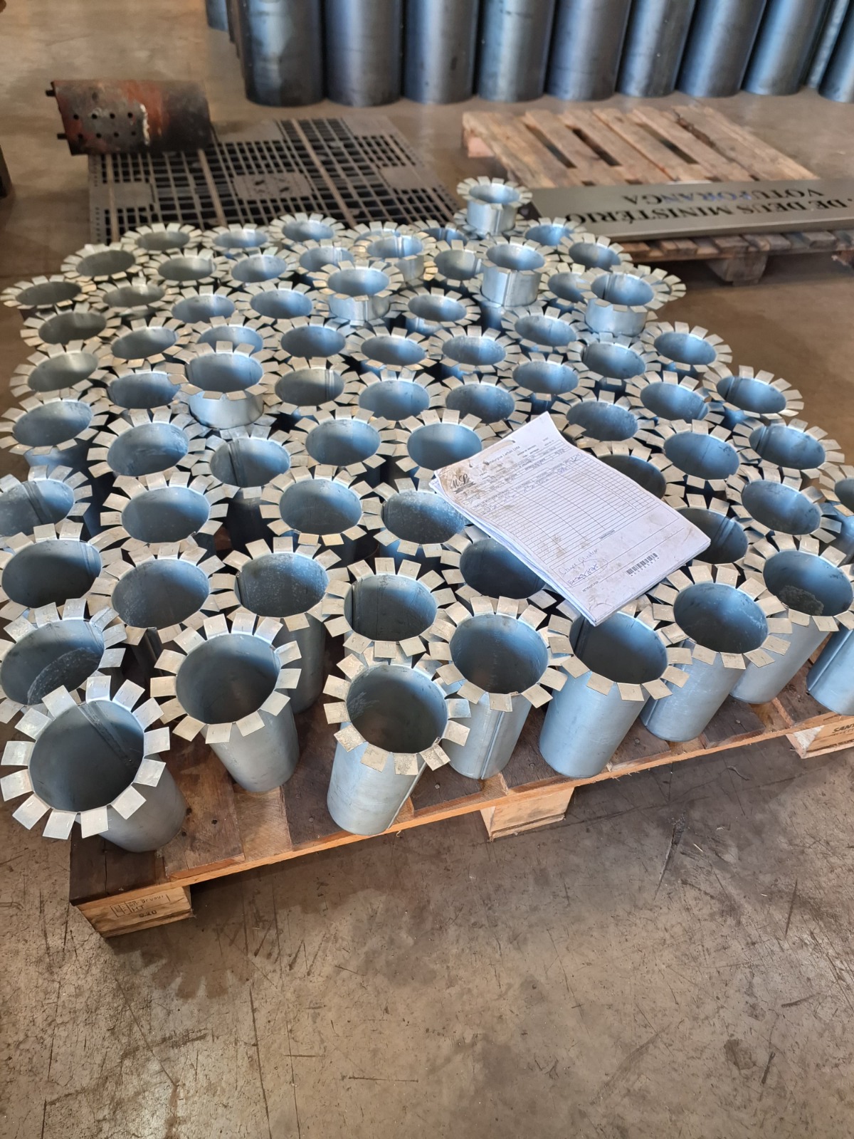 Palete com cilindros metálicos organizados, cada um com abas retangulares no topo, em ambiente industrial com peças e ferramentas ao fundo.