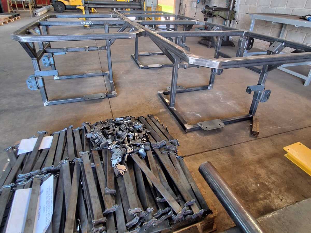 Oficina de fabricação com grandes estruturas metálicas retangulares de tubos soldados, pilha de barras e suportes em palete, e ferramentas ao fundo.