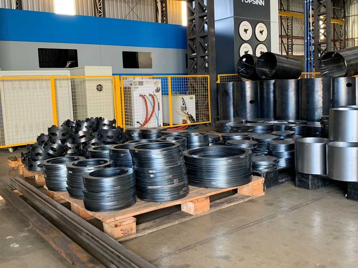 Oficina industrial com peças metálicas organizadas em paletes e no chão, incluindo anéis, cilindros e tubos de aço.