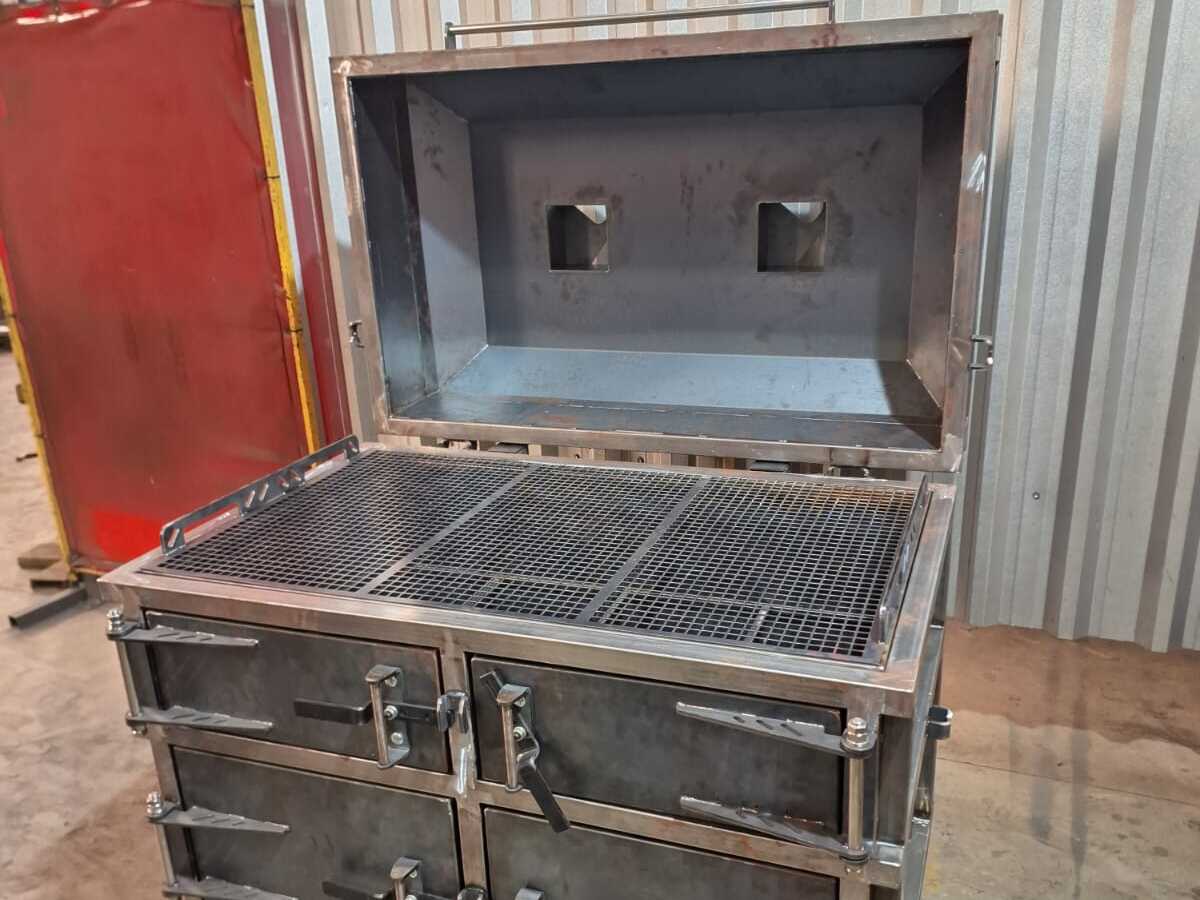Churrasqueira metálica industrial com compartimentos e tampas robustas, em ambiente interno com paredes de metal ondulado.