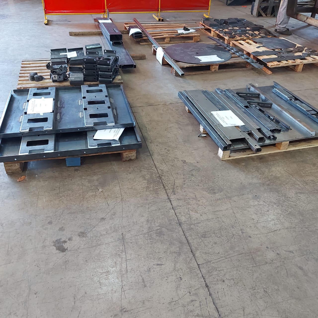 Pallets com peças metálicas variadas, incluindo chapas, vigas e suportes, organizados em chão de oficina industrial.