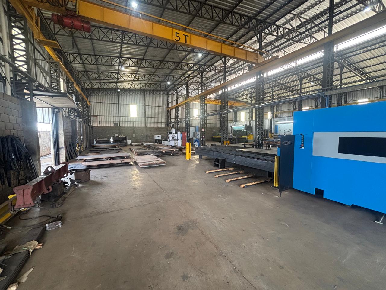Galpão industrial com guindaste aéreo amarelo de 5 toneladas, pilhas de chapas metálicas no chão e máquina azul CNC ao lado.