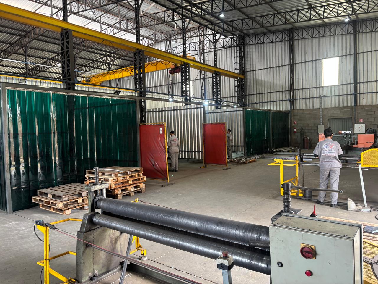 Oficina de metalurgia com máquina de rolagem metálica, cortinas de solda coloridas, guindaste aéreo de 5 toneladas e dois trabalhadores em operação.