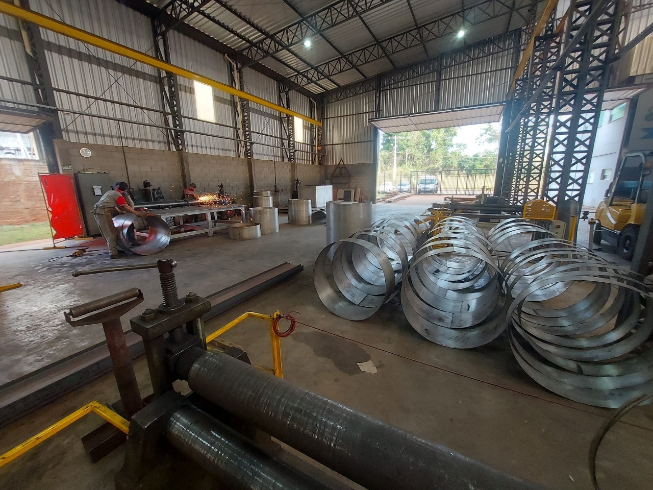 Oficina de metalurgia com máquina de rolagem, grandes anéis metálicos empilhados e trabalhadores usando equipamentos de proteção, um deles produzindo faíscas ao cortar metal.