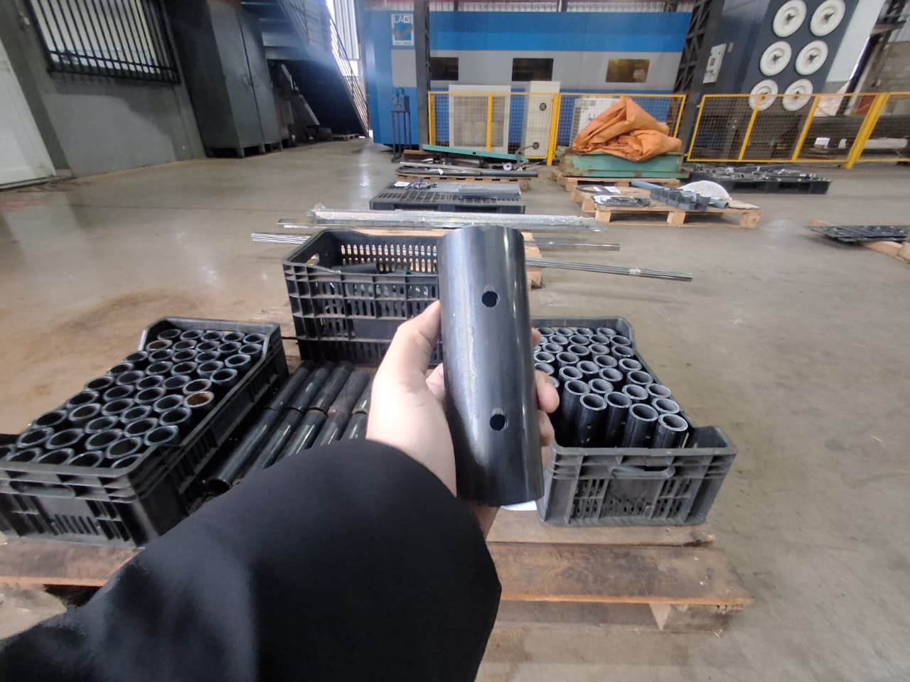 Pessoa segurando tubo metálico com furos laterais em oficina, ao fundo caixas plásticas pretas cheias de peças semelhantes sobre paletes.