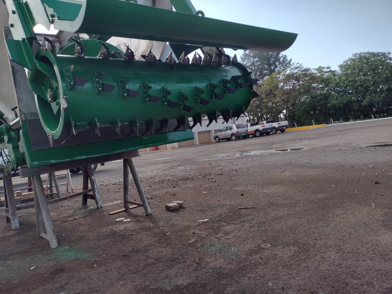 Peça industrial verde em formato cilíndrico com lâminas metálicas afiadas na superfície, montada em estrutura de suporte em área externa.