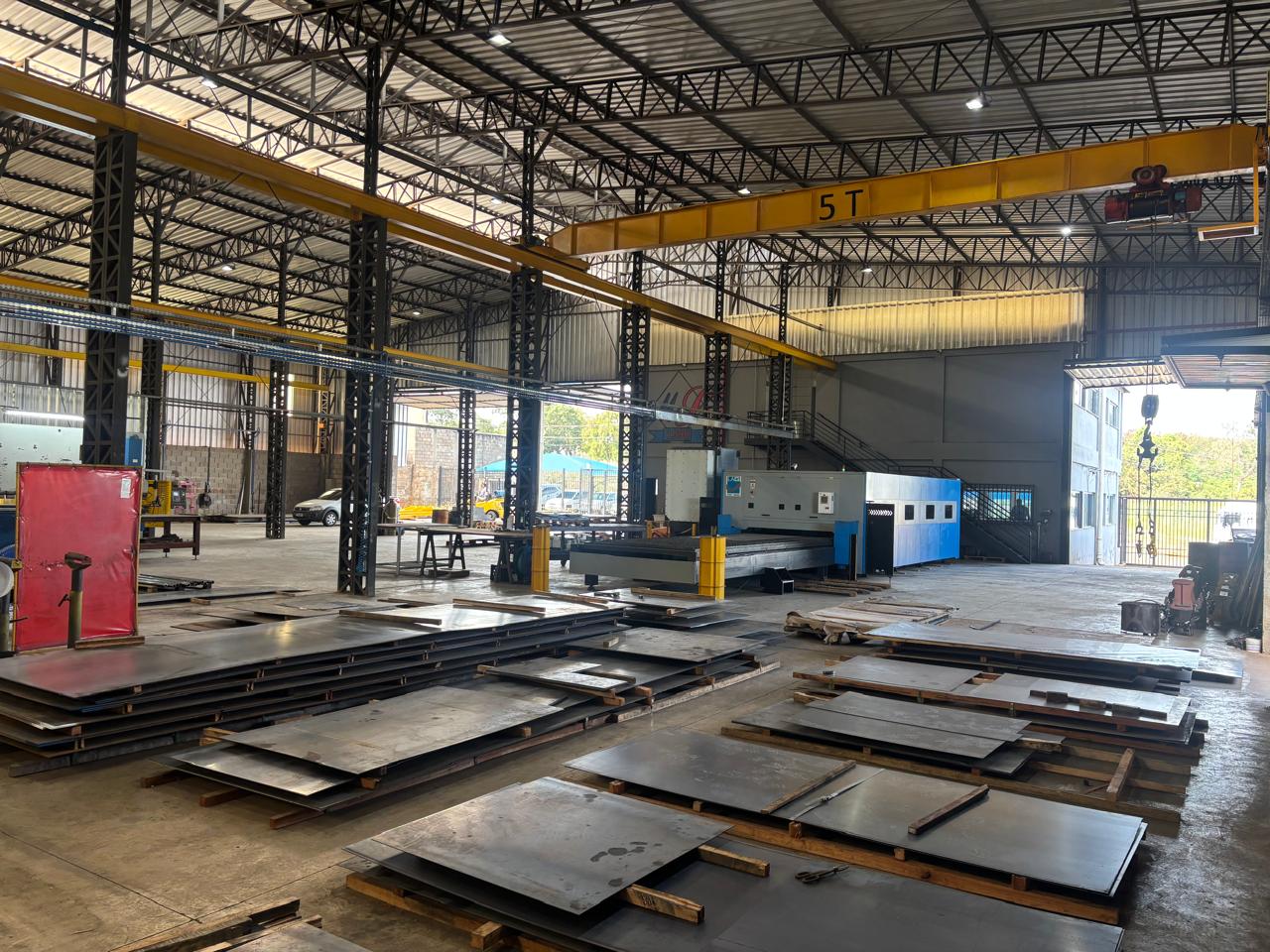 Galpão industrial com chapas metálicas empilhadas em paletes e máquina de corte ao centro.