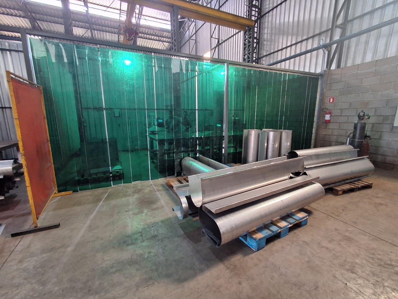 Oficina industrial com grandes componentes cilíndricos metálicos sobre paletes e cortina plástica verde separando área de soldagem.