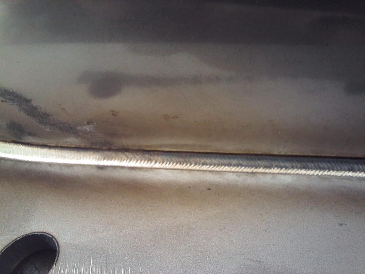 Estrutura metálica com solda horizontal uniforme em superfície de aço, com marcas de calor e furo circular visível.