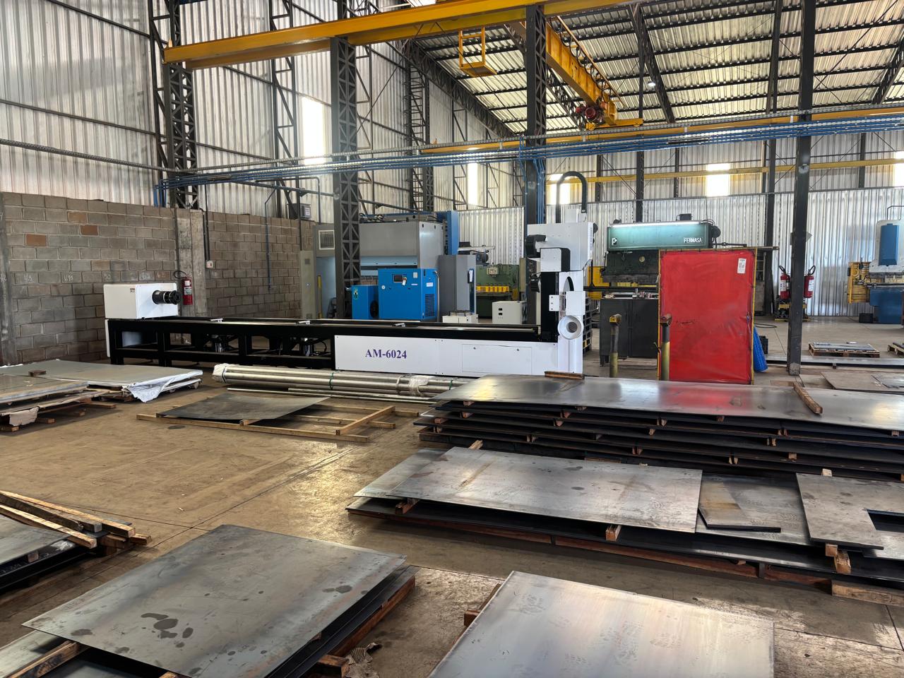 Oficina industrial com máquina CNC ao centro, chapas metálicas empilhadas em paletes e outras máquinas ao fundo.