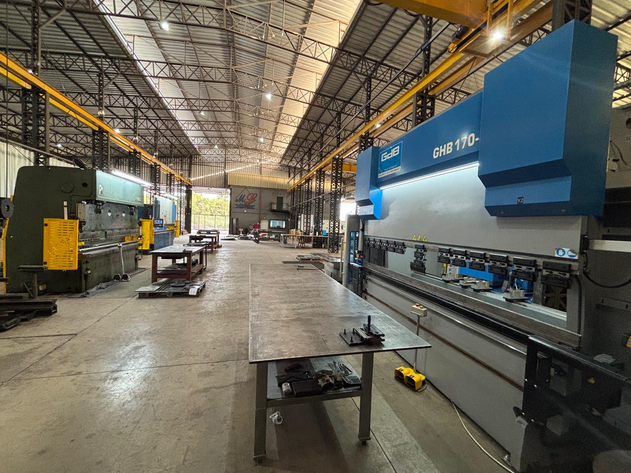 Oficina industrial ampla com prensas dobradeiras verde e azul, mesas de trabalho centrais e guindastes amarelos suspensos.
