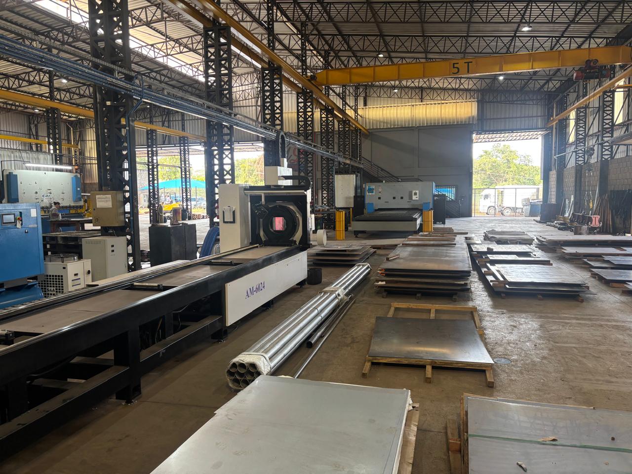 Oficina industrial ampla com máquinas pesadas, chapas e barras metálicas organizadas no chão e guindaste aéreo amarelo de 5 toneladas.