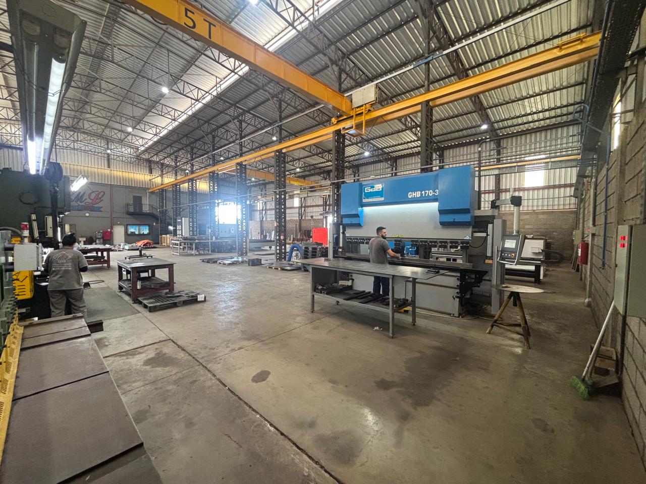 Galpão industrial com máquinas pesadas, guindaste aéreo de 5 toneladas e dois trabalhadores operando equipamentos.