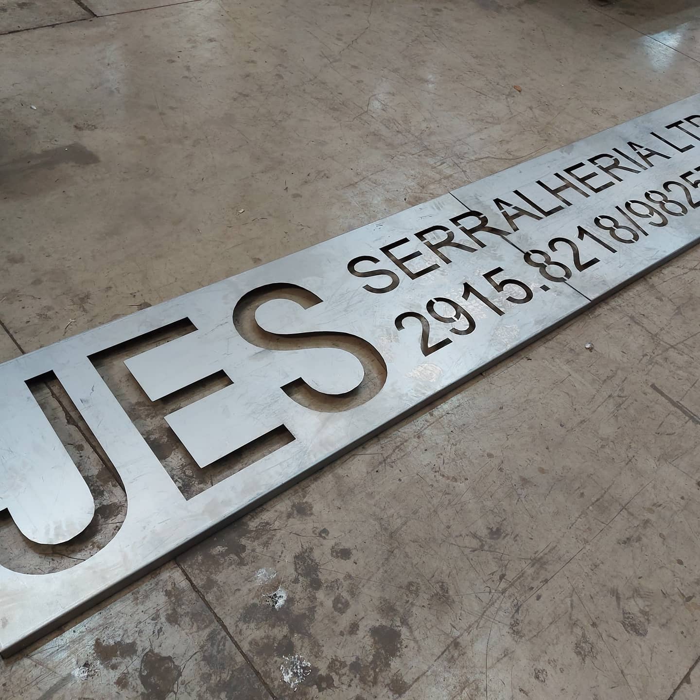 Placa metálica com letras e números recortados, posicionada no chão de oficina.