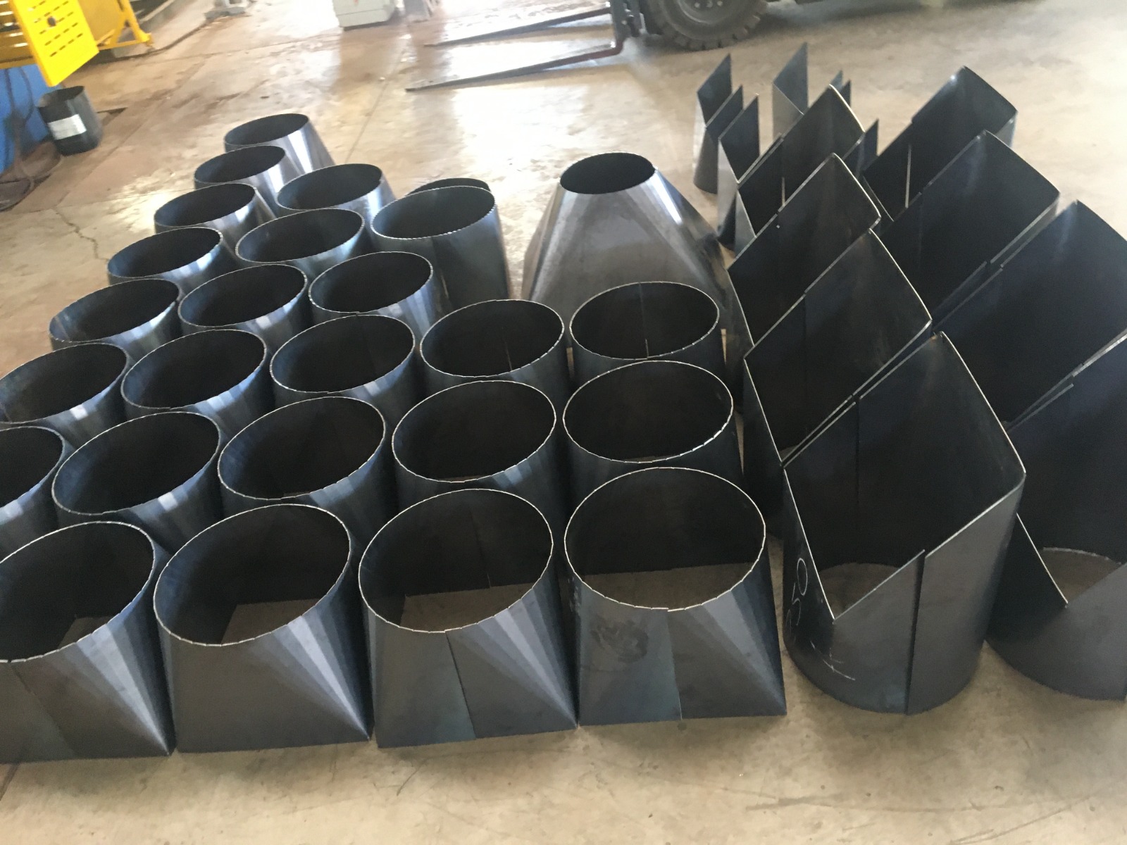 Componentes metálicos cilíndricos e cônicos organizados no chão de oficina industrial, e equipamentos ao fundo.