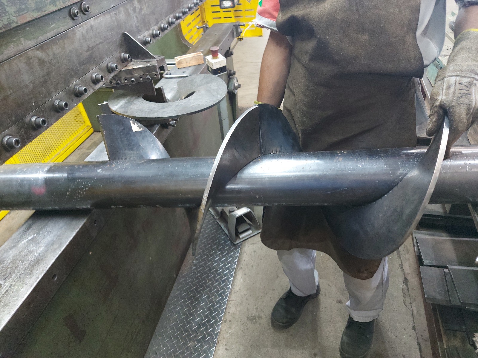 Trabalhador em oficina segurando eixo metálico com hélice soldada, cercado por bancada e equipamentos industriais.