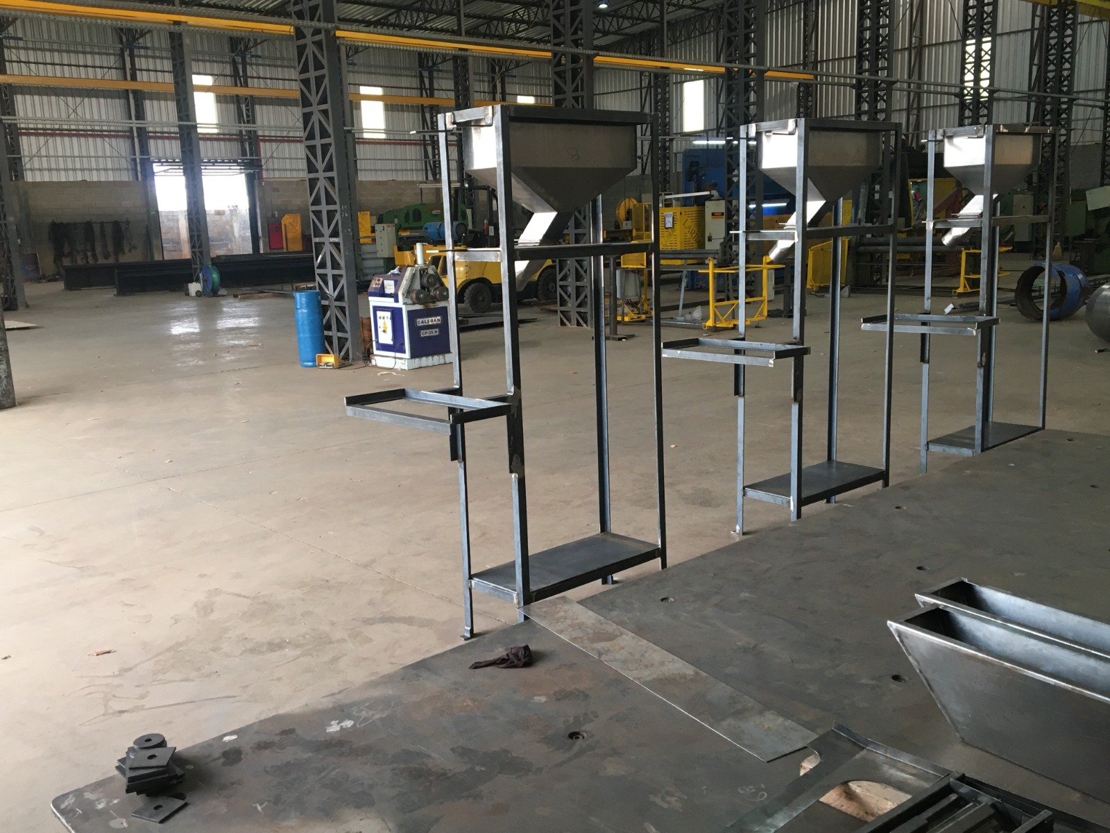 Interior de fábrica com estruturas metálicas em formato de funil alinhadas, empilhadeiras e equipamentos industriais ao fundo.