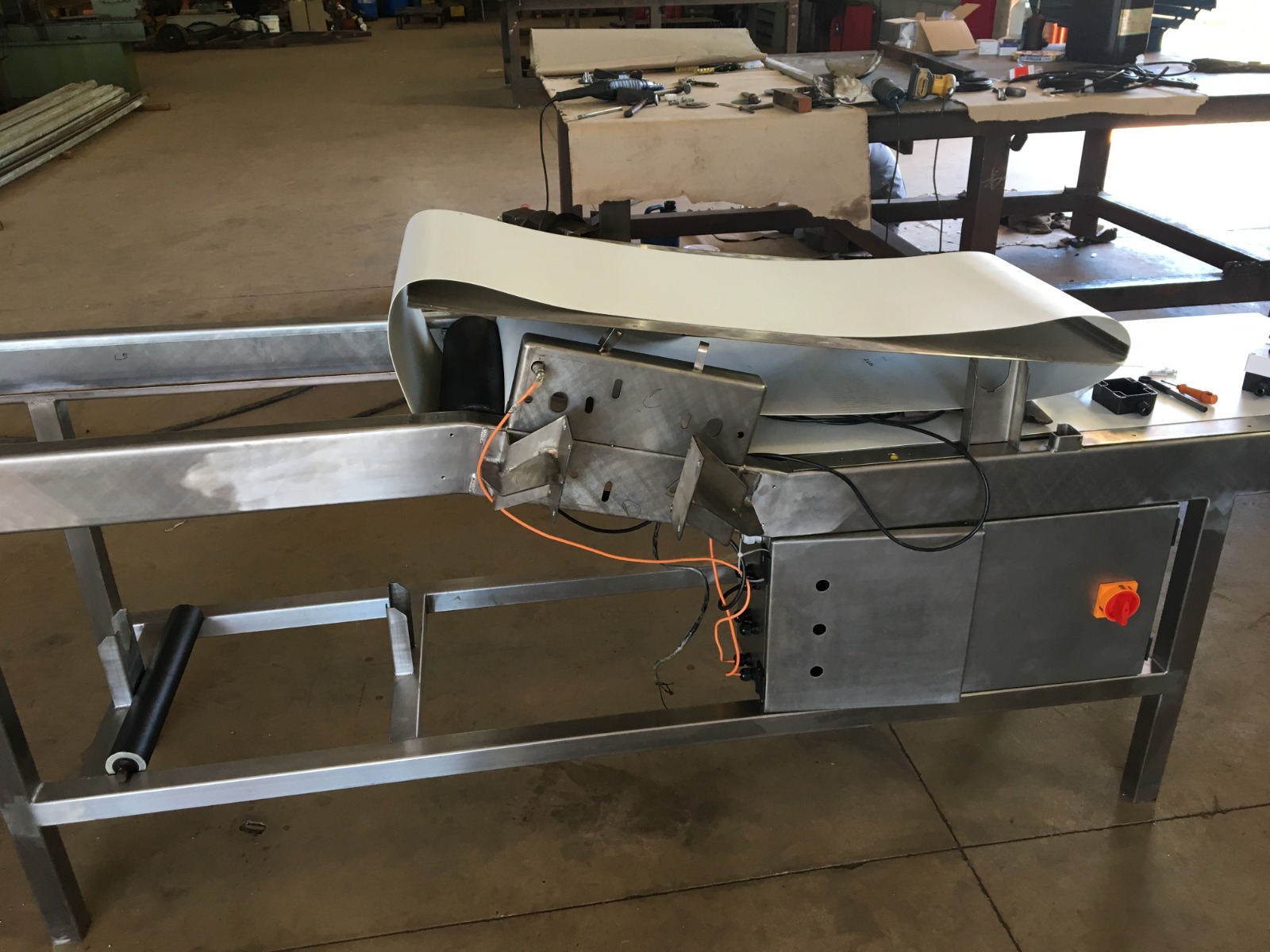 Esteira transportadora industrial parcialmente montada em aço inox, com correia branca e caixa de controle lateral em oficina.