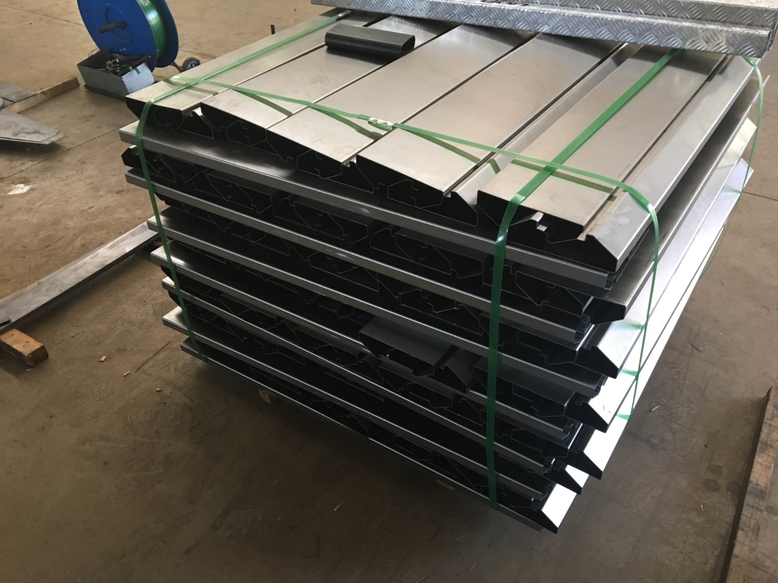 Pilhas de perfis metálicos dobrados e amarrados com fitas verdes sobre palete de madeira em oficina industrial.