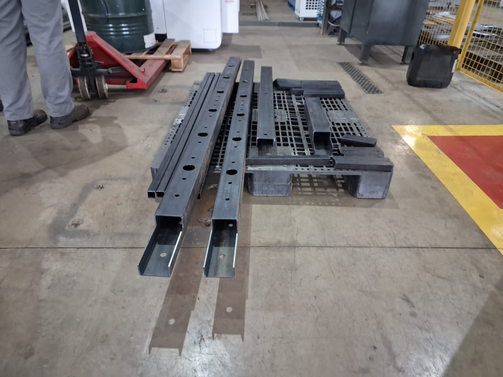 Vigas de aço retangulares com furos circulares empilhadas em palete, ao lado de operador com paleteira manual em oficina industrial.