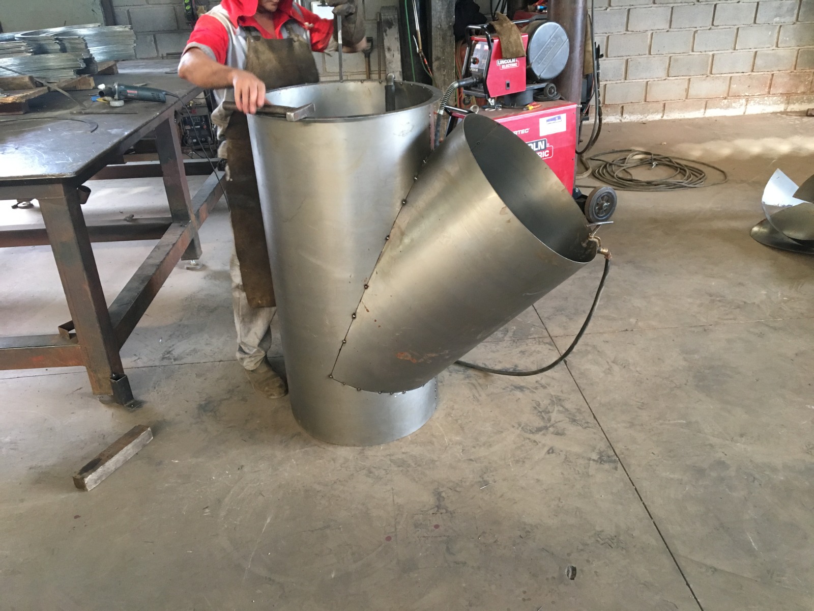 Trabalhador em oficina de metalurgia manuseando peça cilíndrica metálica com seção cônica soldada, cercado por bancada, máquina de solda e componentes industriais.