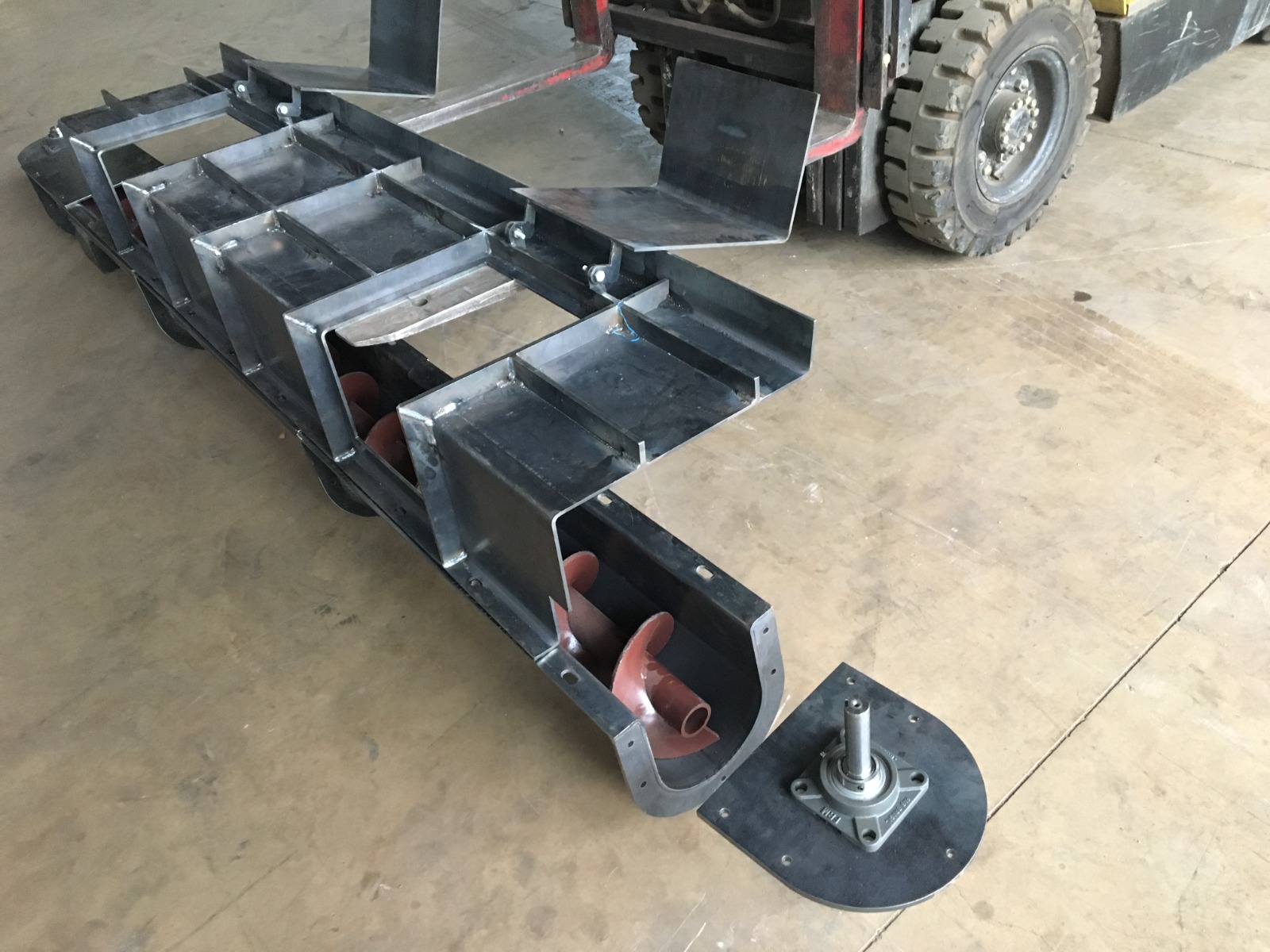 Montagem de rosca transportadora vermelha em estrutura metálica, posicionada no chão de oficina industrial próxima a empilhadeira.