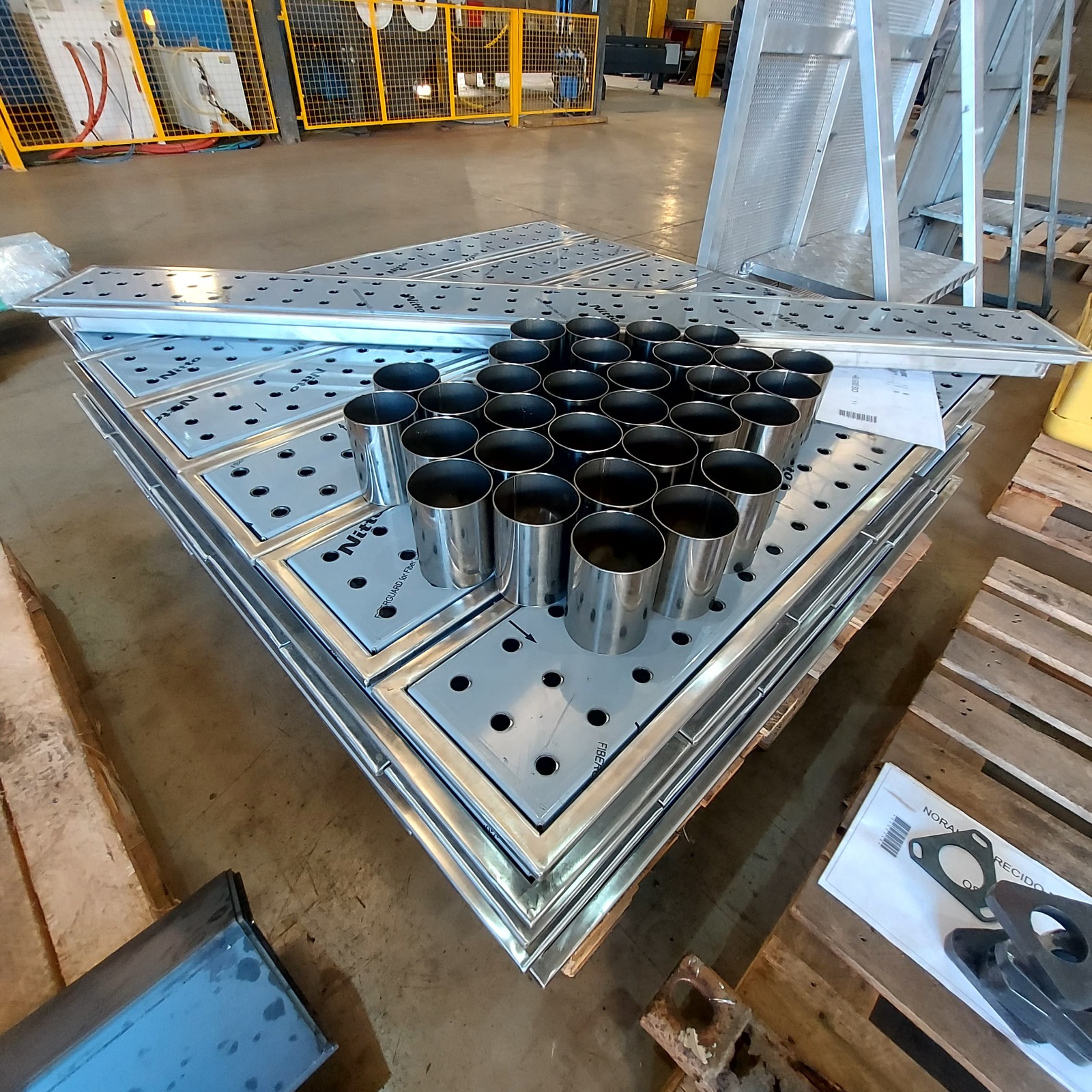 Palete com placas metálicas perfuradas e tubos cilíndricos organizados em oficina industrial.