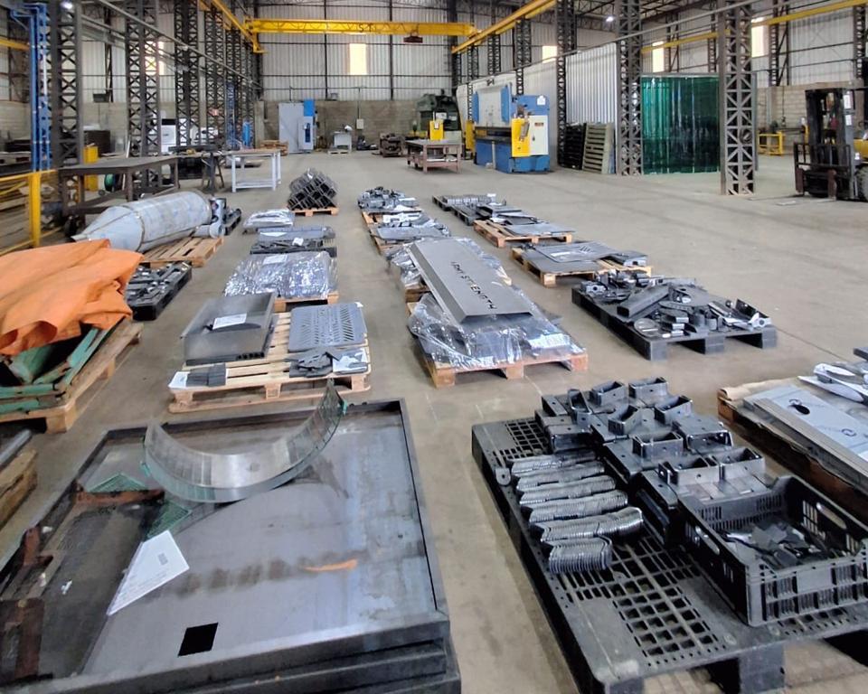 Oficina industrial com peças metálicas variadas organizadas em paletes e caixas, máquinas pesadas e ponte rolante ao fundo.