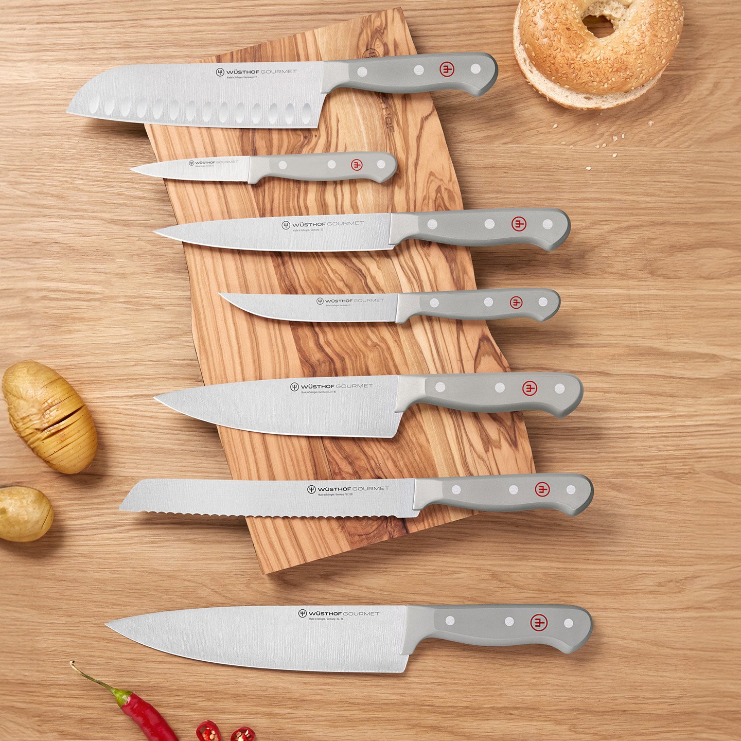 a variety of grey knives sitting atop of wood cutting board with various pieces of food surrounding