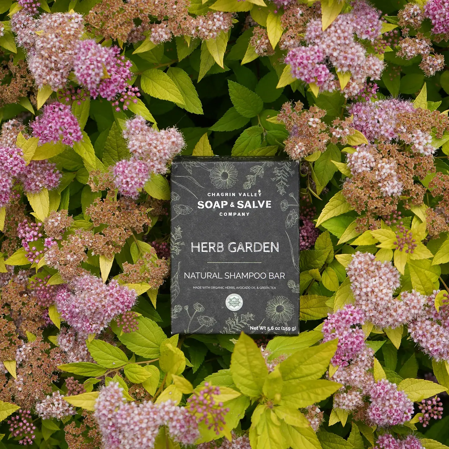 Natural shampoo bar labeled "Herb Garden" by Chagrin Valley Soap & Salve, surrounded by lush green leaves and pink flowers.