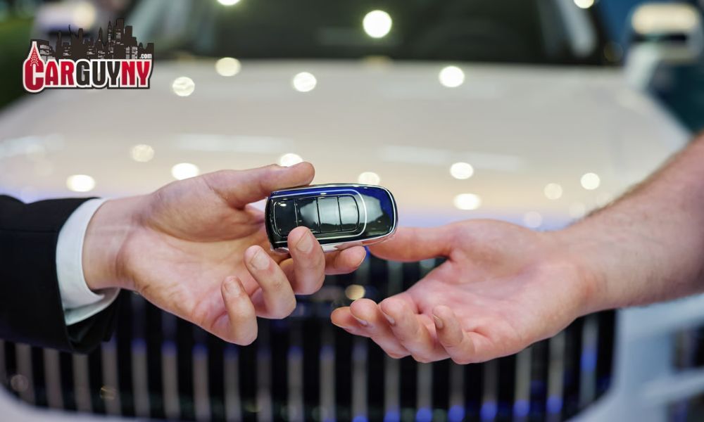Hand holding a car key fob passing it to another hand, with a car grill in the background. Logo reading "CarGuyNY" on the top left.