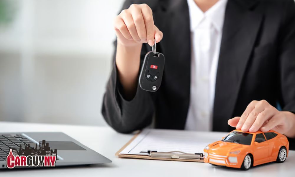 Person in a suit holding car keys above a clipboard, with a laptop and toy car on the desk.