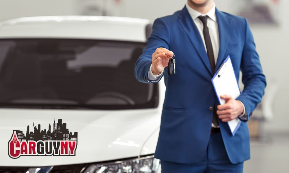 Man in a blue suit holding a car key and clipboard, standing in front of a white car with "CarGuyNY.com" logo.