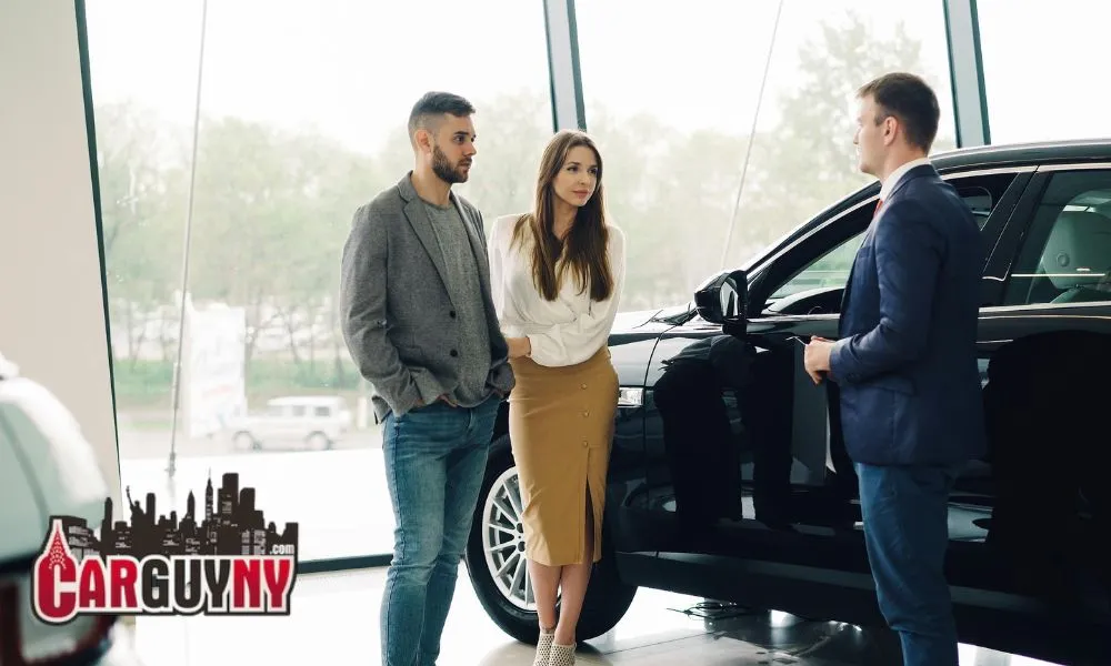 A couple converses with a salesman next to a black car in a showroom, with large windows in the background and a "Car Guy NY" logo visible.