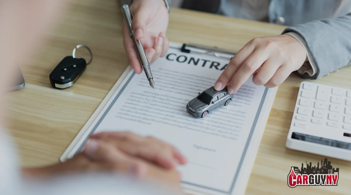 Hands signing a car contract with a small car model on the document. Car keys and a calculator are on the wooden table.