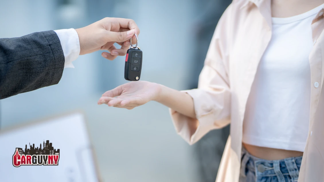 A person hands over car keys to another outside, with a blurred "CarGuyNY" logo visible in the foreground.