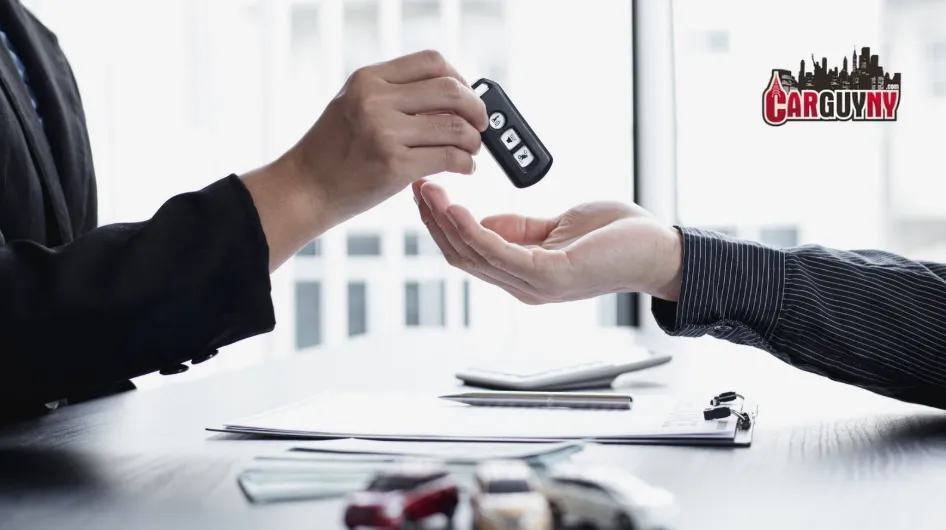 Person handing over car keys to another person across a table with documents, calculator, and toy cars; "Car Guy NY" logo in the background.
