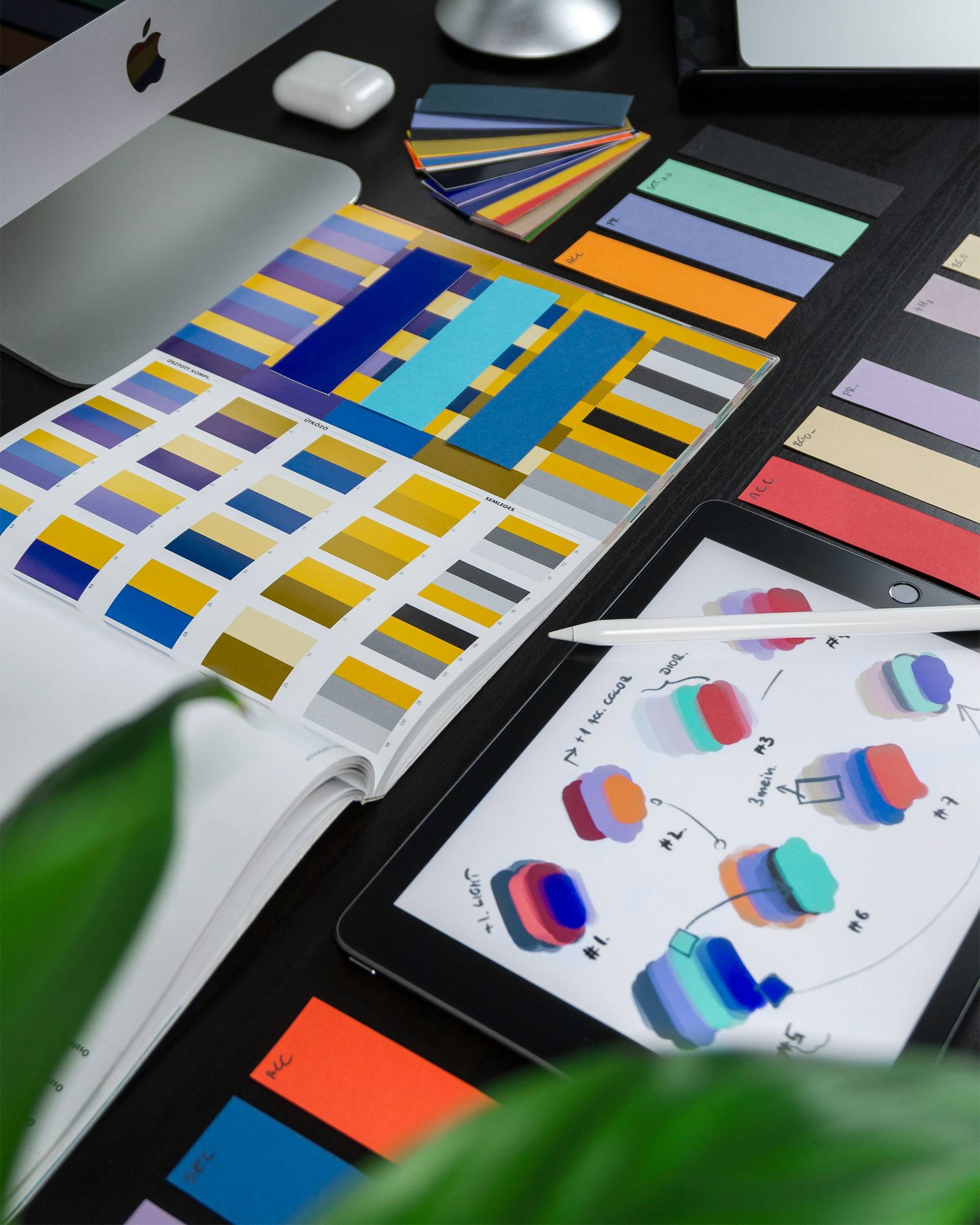 A workspace with color swatches, an iPad displaying layered designs, and a stylus on a dark desk, partially obscured by a plant.