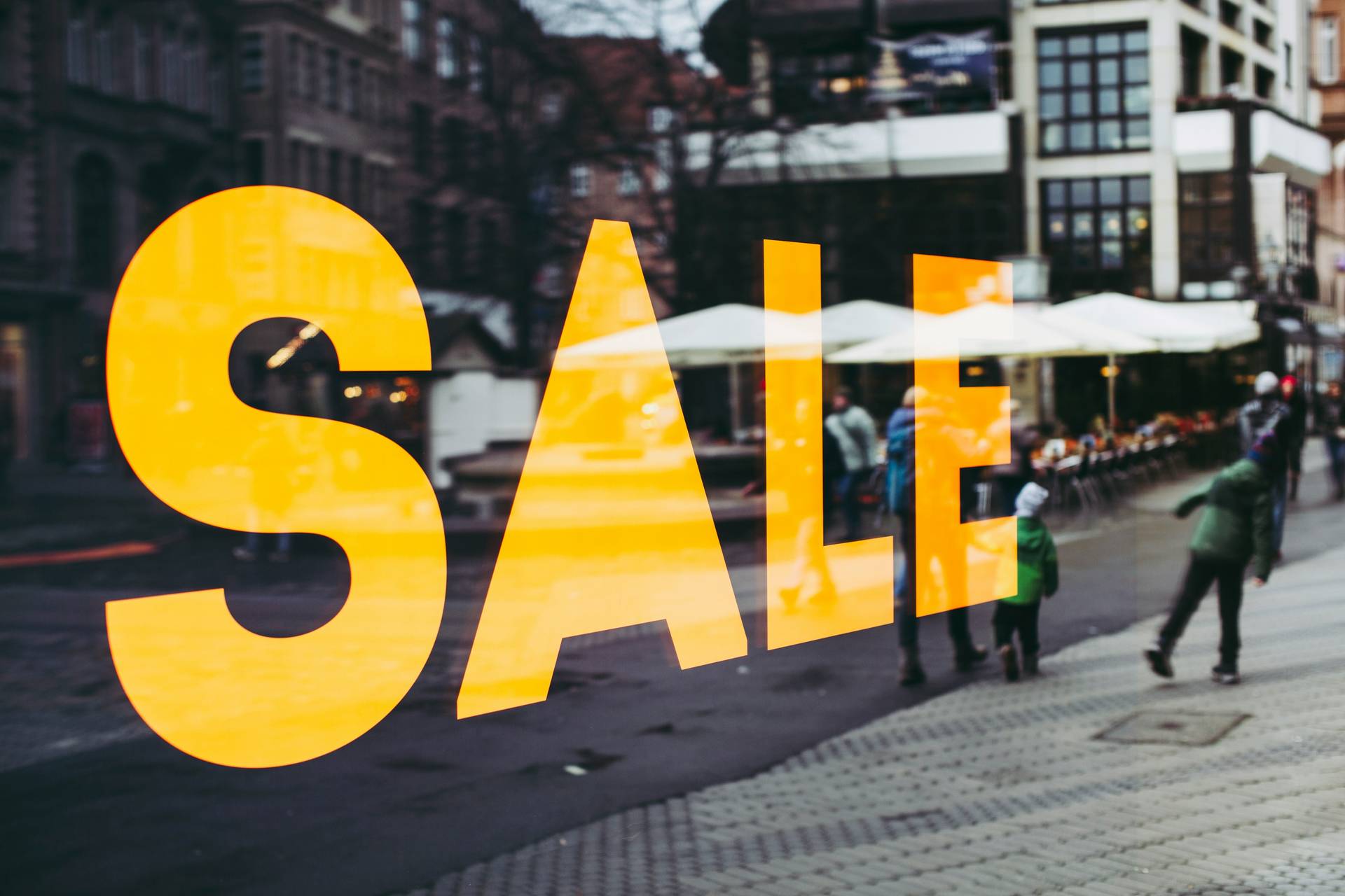 Large yellow "SALE" sign on a storefront window, reflecting a busy street scene with people walking and outdoor seating.