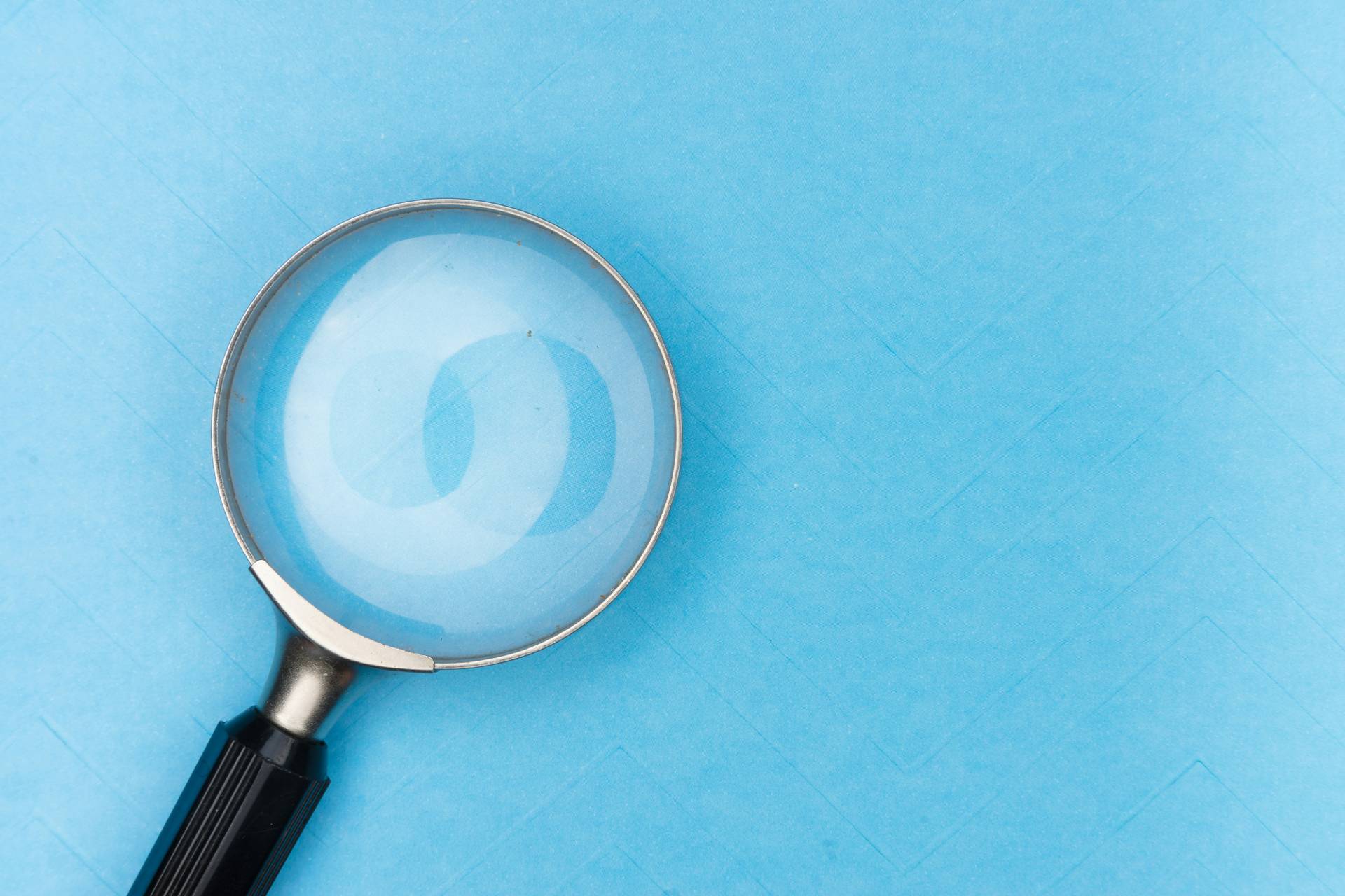 A magnifying glass with a black handle lies on a light blue surface, casting a shadow and slightly magnifying the background pattern.
