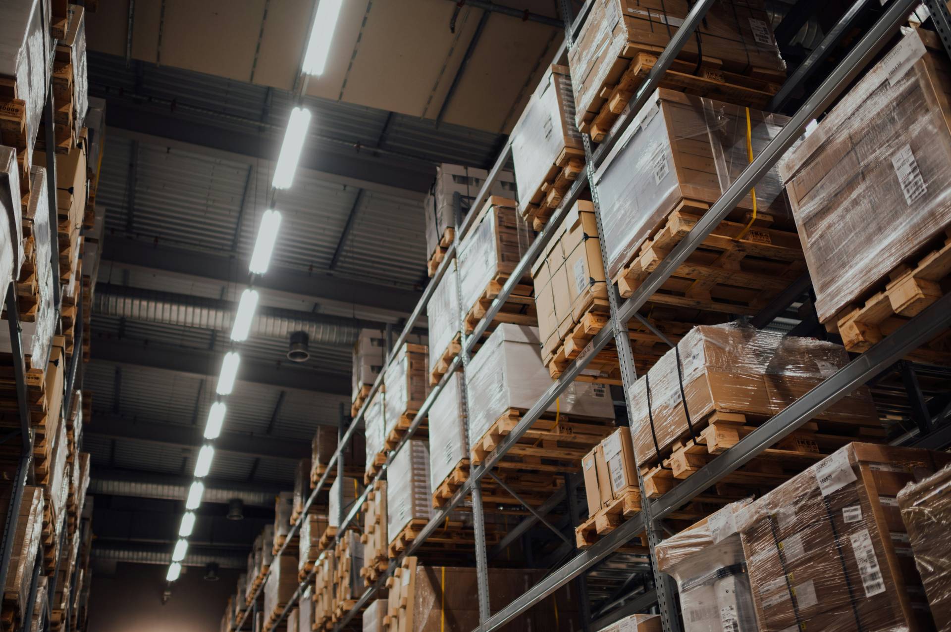 Warehouse interior with tall shelves filled with stacked pallets of boxes, under bright overhead lighting.