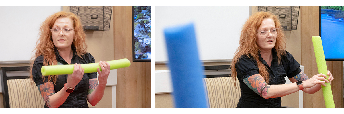 A person with long red hair holds and gestures with a green foam pool noodle during a presentation indoors.