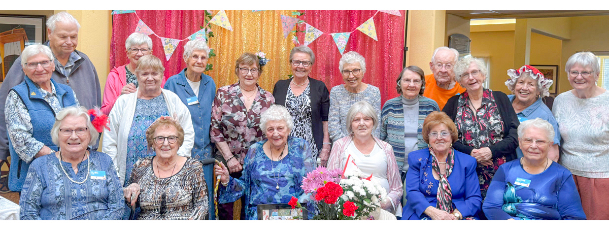 A group of smiling elderly people, mostly women, dressed in colorful clothing, posing together in front of a bright, decorated background.
