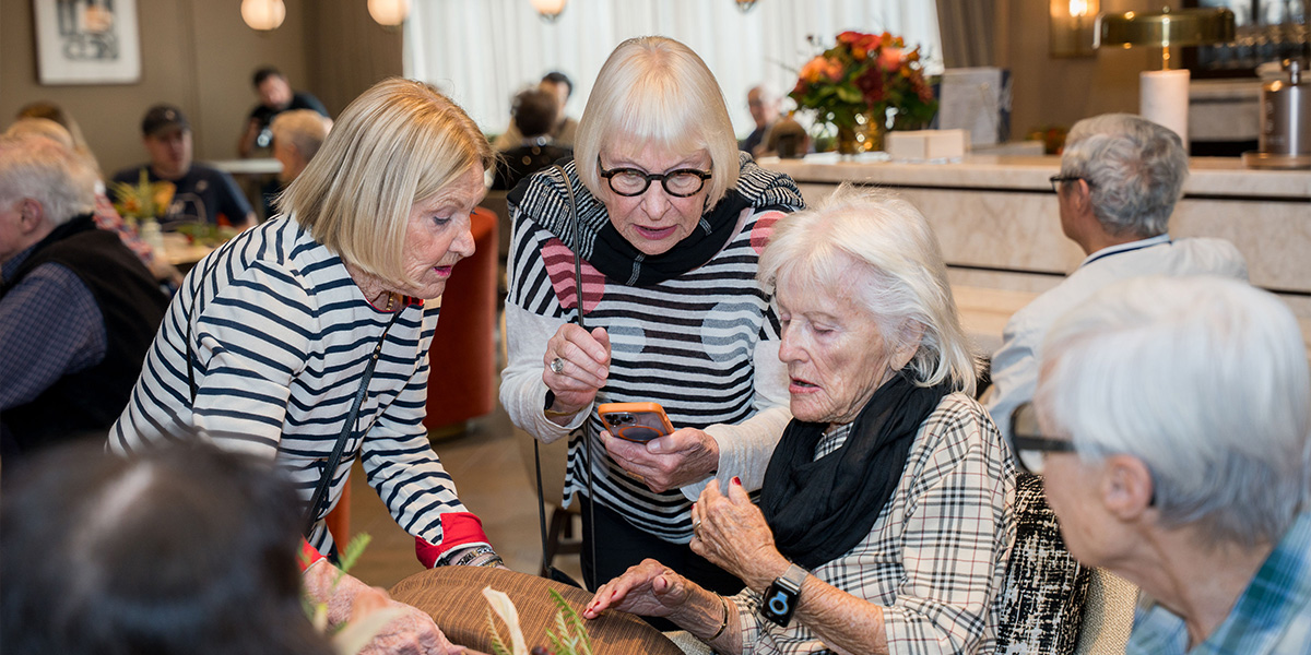 Group of senior women gathered at a table, engaged in conversation, with one showing a smartphone. Others are seated in the background.