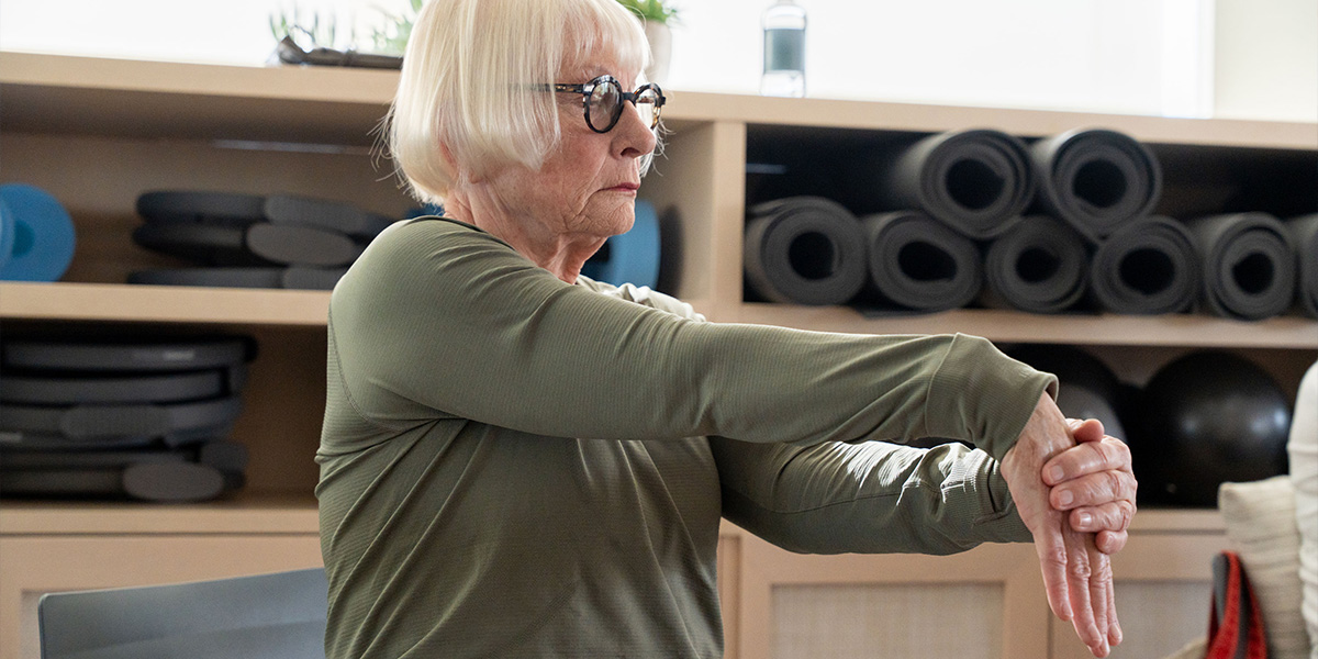 Carol, wearing a green top, stretches her arms in a gym, with shelves of yoga mats and equipment in the background.
