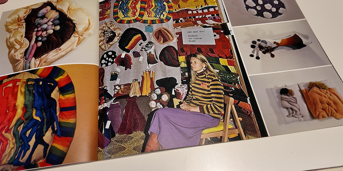 Carol, in a striped outfit, sits in a colorful, textile-filled room with vibrant patterns and yarn art on the walls and floor.