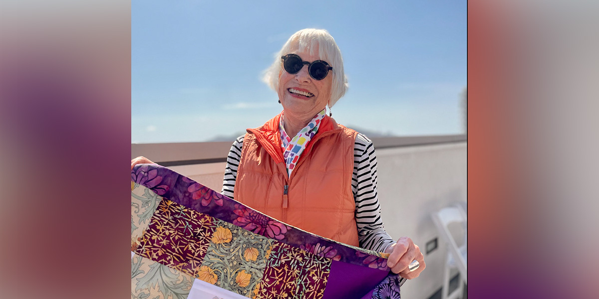 Carol, in sunglasses and an orange vest, holds a colorful quilt, smiling outdoors on a sunny day.