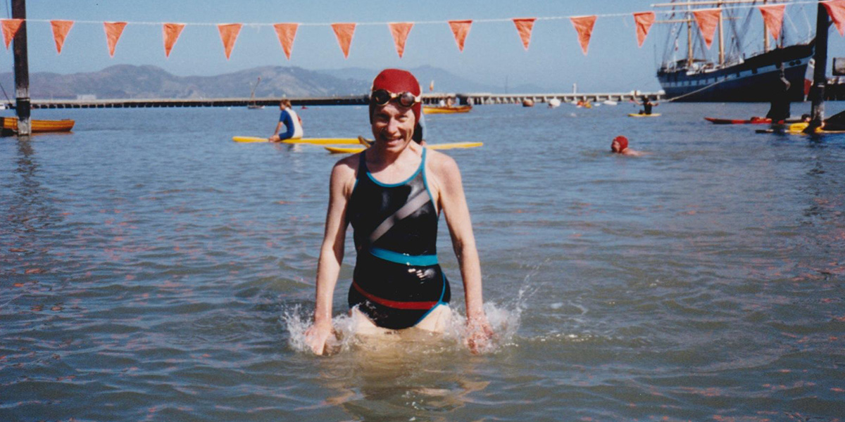 Carol wearing a black swimsuit and red swim cap stands in the water, smiling. Sailboats and mountains are visible in the background.